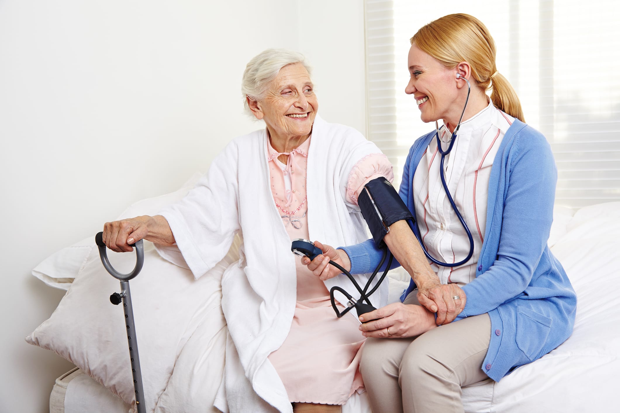 A carer checking an older woman's blood pressure as they sit on a bed