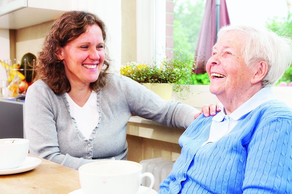 A carer and an older woman laughing together at a kitchen table