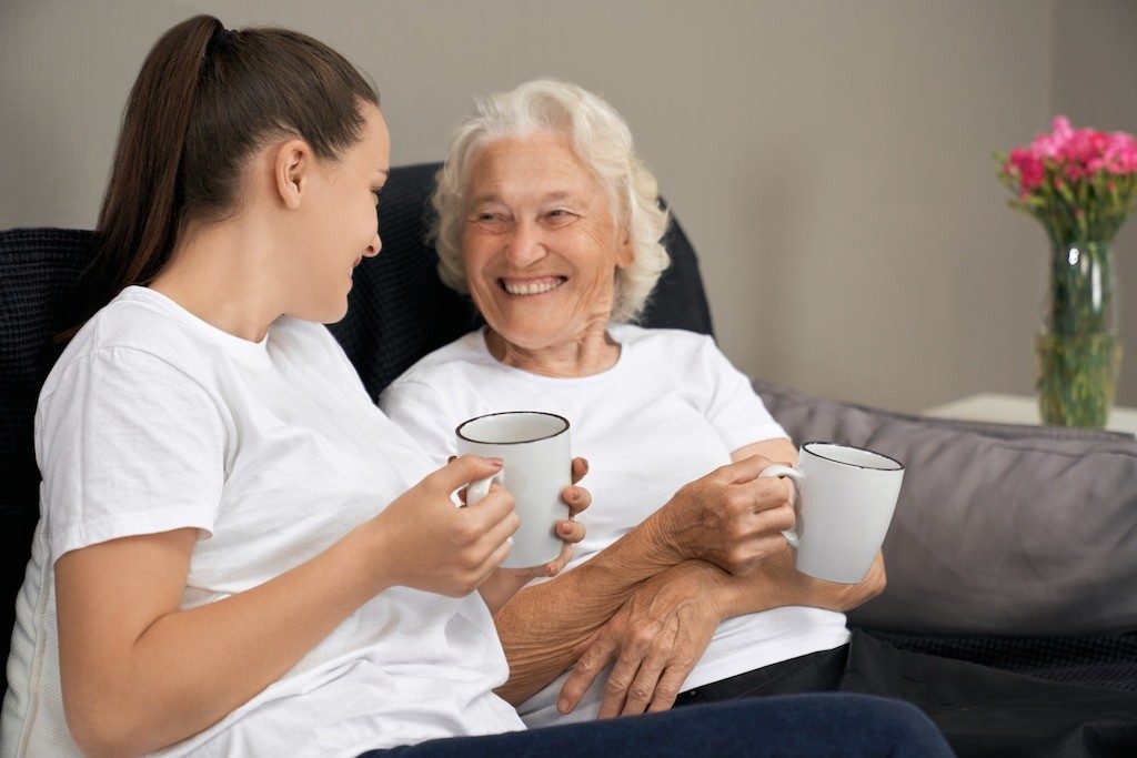 A carer and an older woman sharing mugs of tea on the sofa