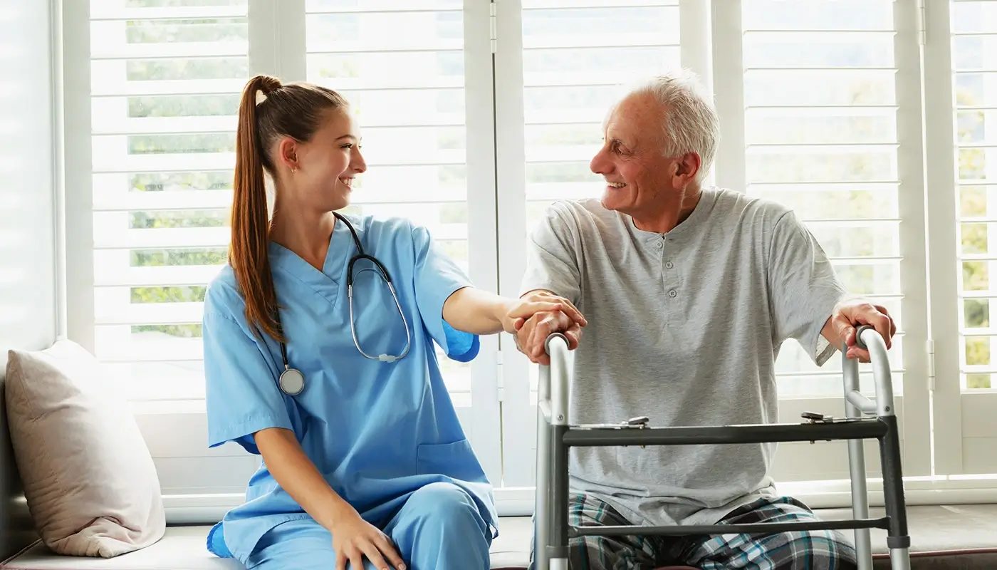 A nurse sitting with an older man who has a walking frame beside a window