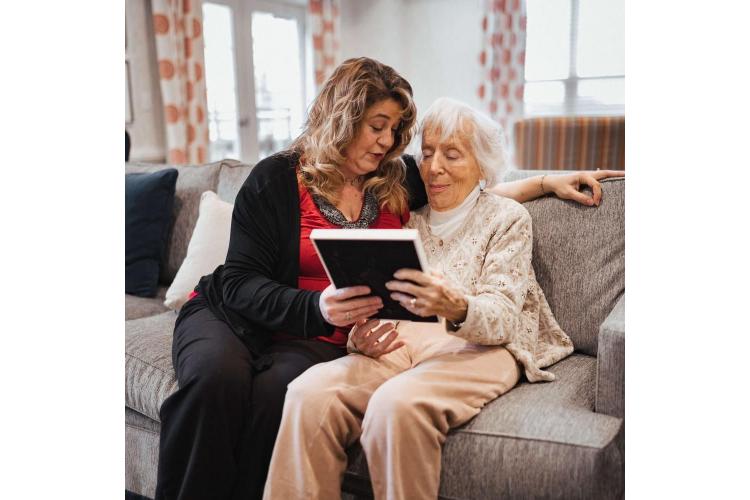 A carer and an elderly woman looking at a tablet together on the sofa
